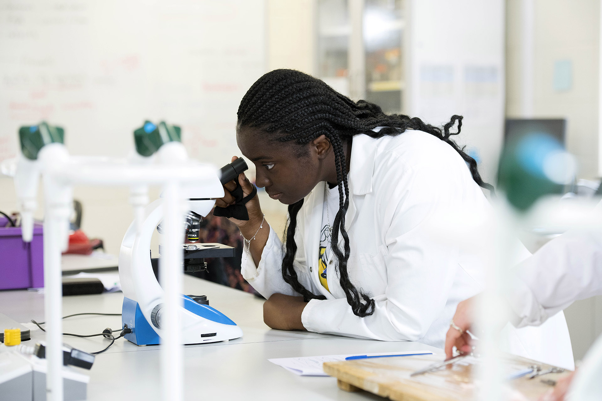 A woman in a lab coat examines a sample under a microscope in a laboratory setting, surrounded by equipment and notes.